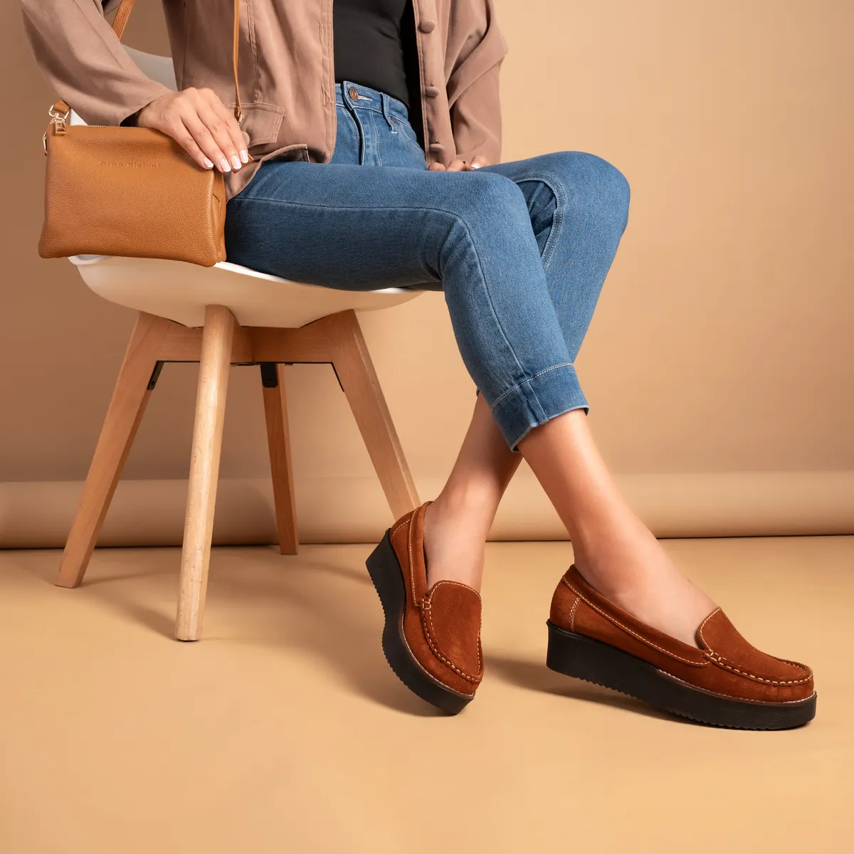 A woman in denim trying on tan loafers, seated on a wooden chair