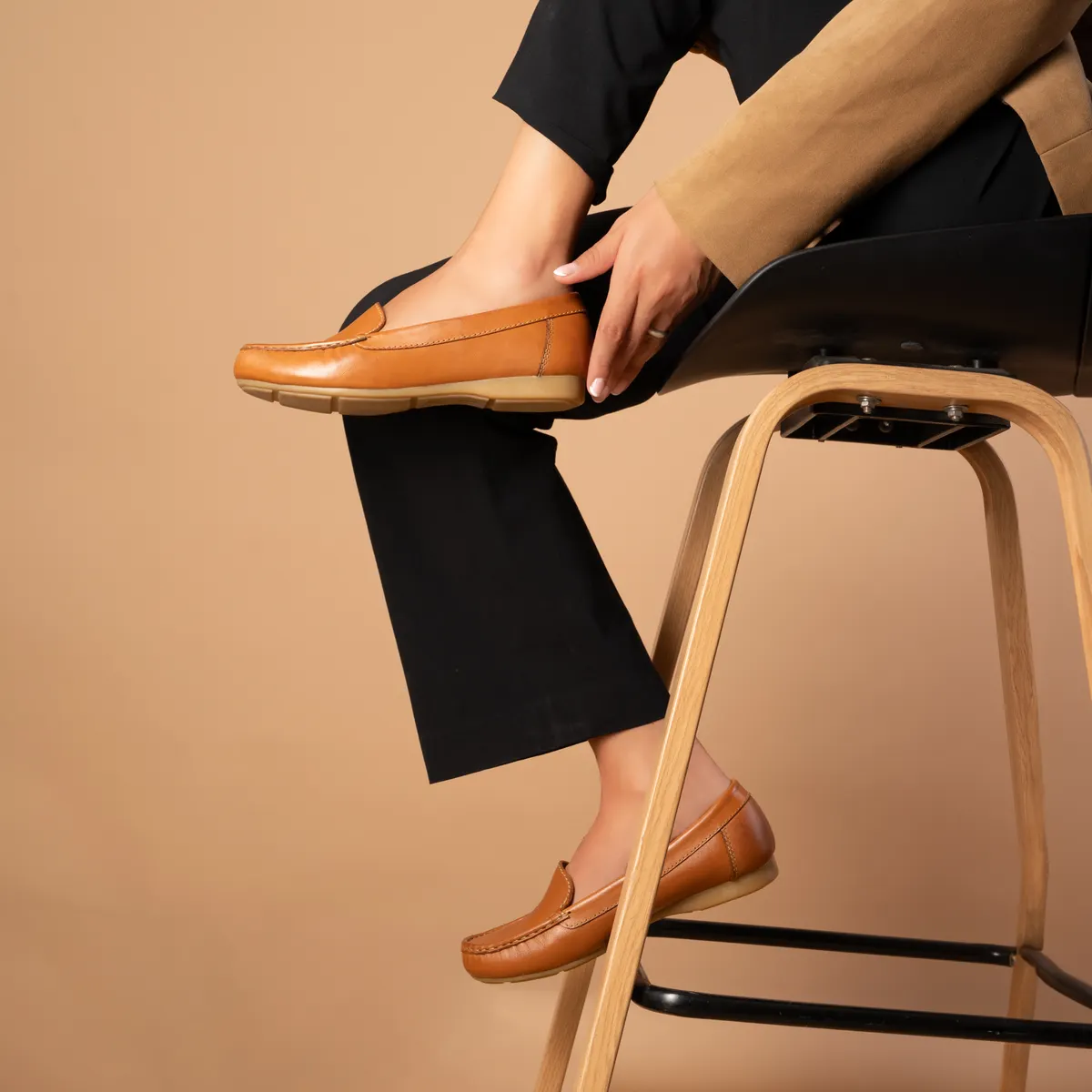Tan suede loafers worn with black trousers on a wooden stool